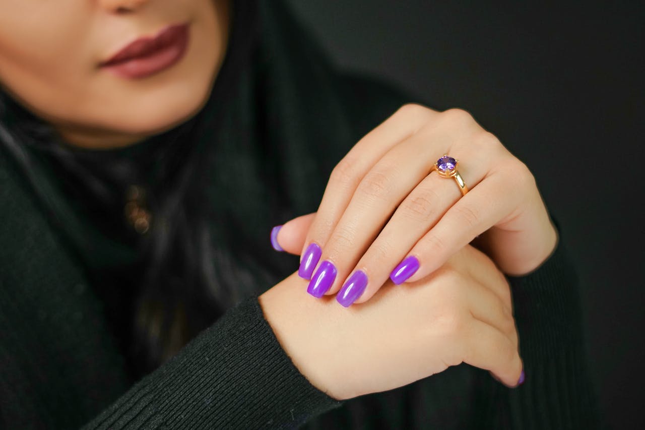 hero-img-01 Close-up of woman's hands with vibrant purple nails and a gold ring, set against a dark background.